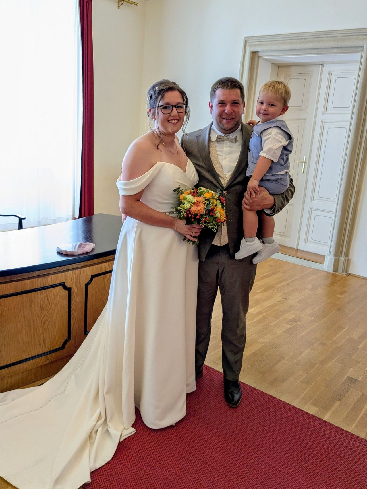 Ein frisch verheiratetes Paar mit ihrem kleinen Sohn, die Braut in einem weißen Kleid, der Bräutigam in einem Anzug und der Junge in einem blauen Outfit, stehen in einem Raum mit Holzböden und einem roten Teppich.