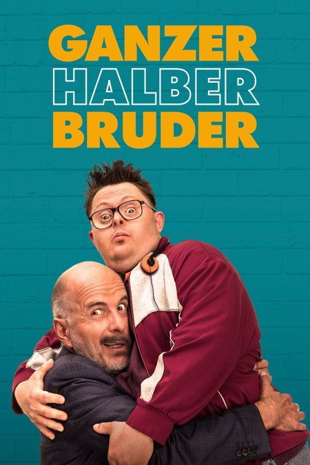 Zwei Männer, einer mit Brille und einer burgunderroten Jacke, der andere kahlköpfig und in einem Anzug, umarmen sich vor einer türkisfarbenen Ziegelwand mit dem Schriftzug 'Ganzer Halber Bruder' in Gelb.