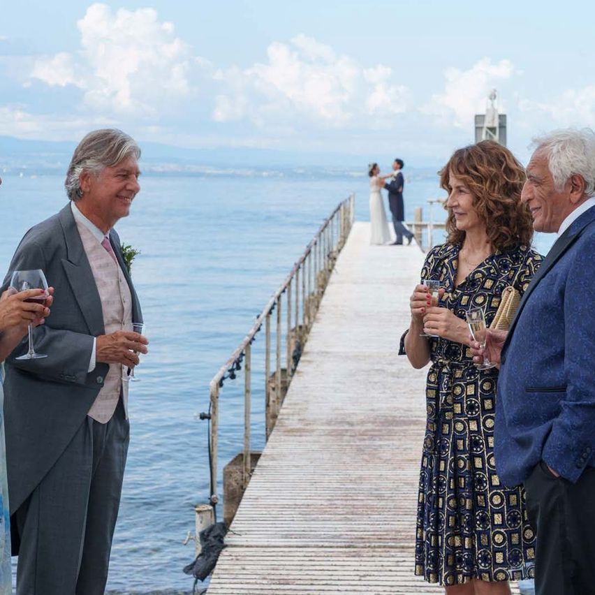 Eine Gruppe von Menschen in formeller Kleidung steht auf einem Pier, hält Weingläser in der Hand und blickt auf das Wasser. Im Hintergrund tanzt ein Paar auf dem Pier.