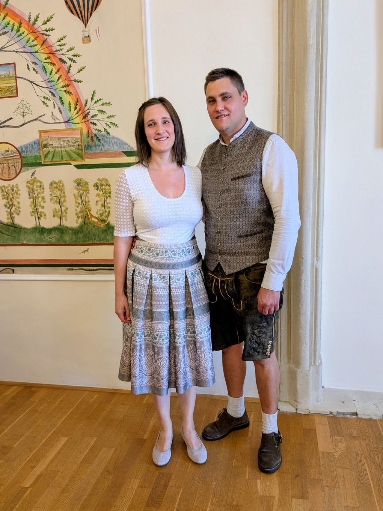 A man and a woman stand close together in a room with a mural behind them. The woman wears a white blouse and a gray skirt. The man wears a vest over a white shirt and black shorts.