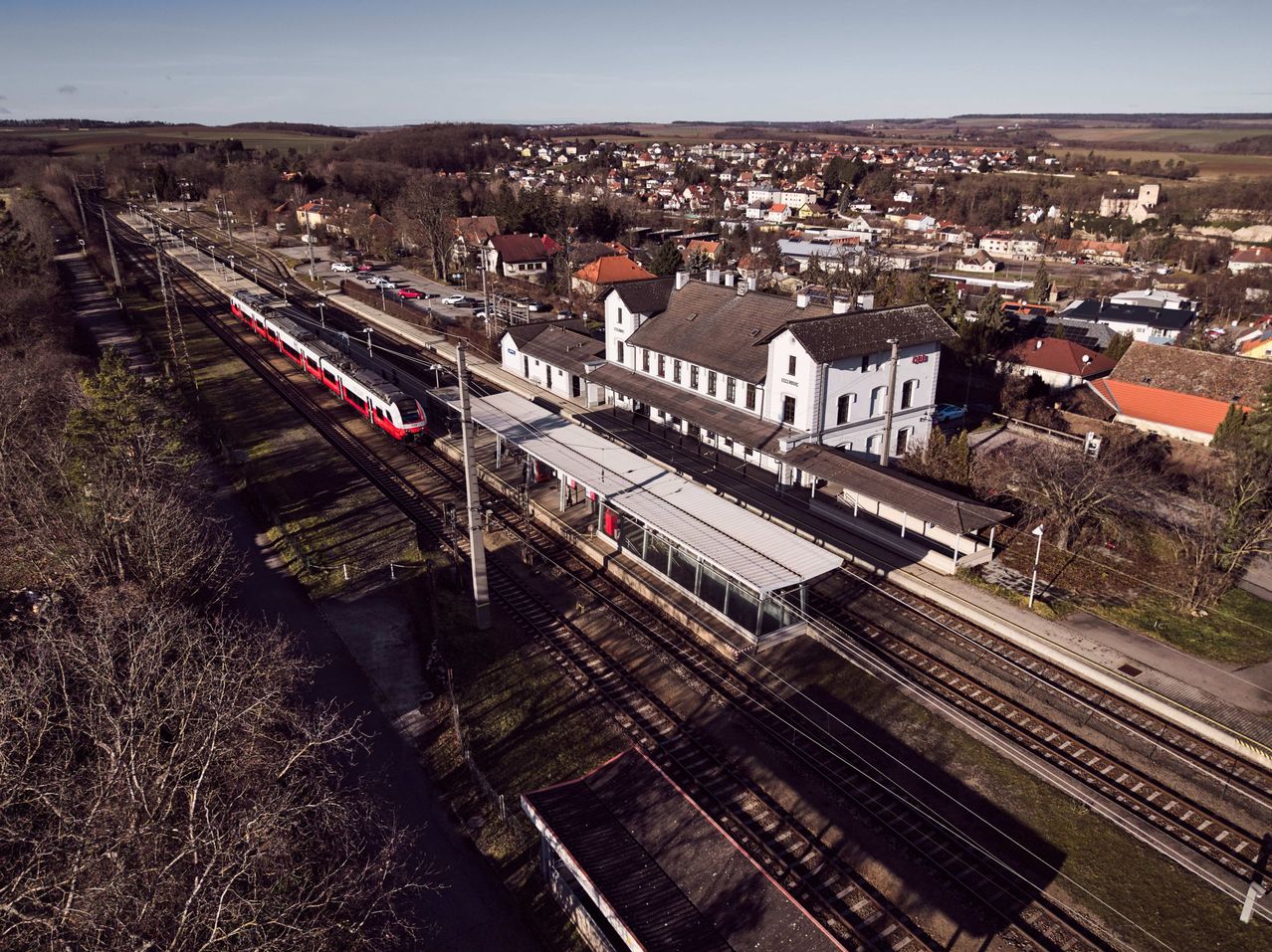 Aerial view of a train station with a red and white train on the tracks, a large white building, and a shelter.