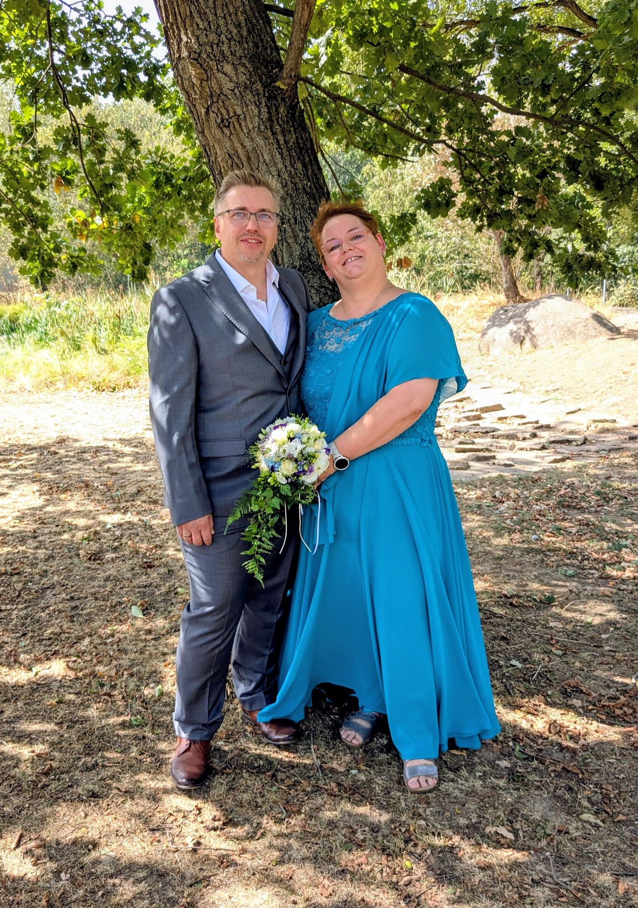 A man and a woman stand together under a tree, smiling for a photo. The woman holds a bouquet of flowers.
