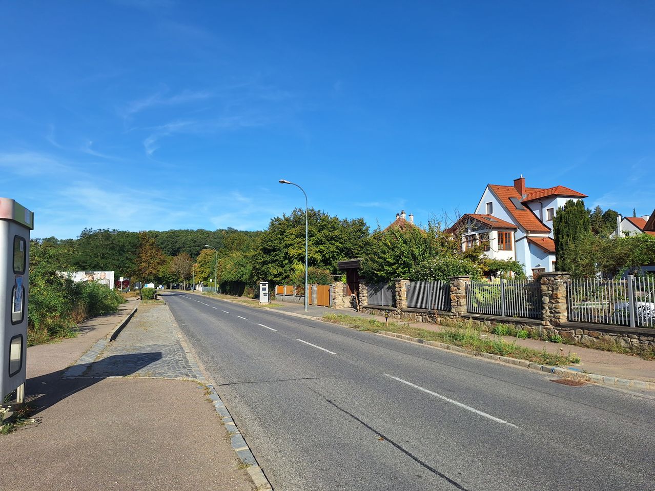 An empty street with a clear blue sky, lined with houses and trees on either side. The houses have red roofs and are surrounded by stone walls and metal fences.