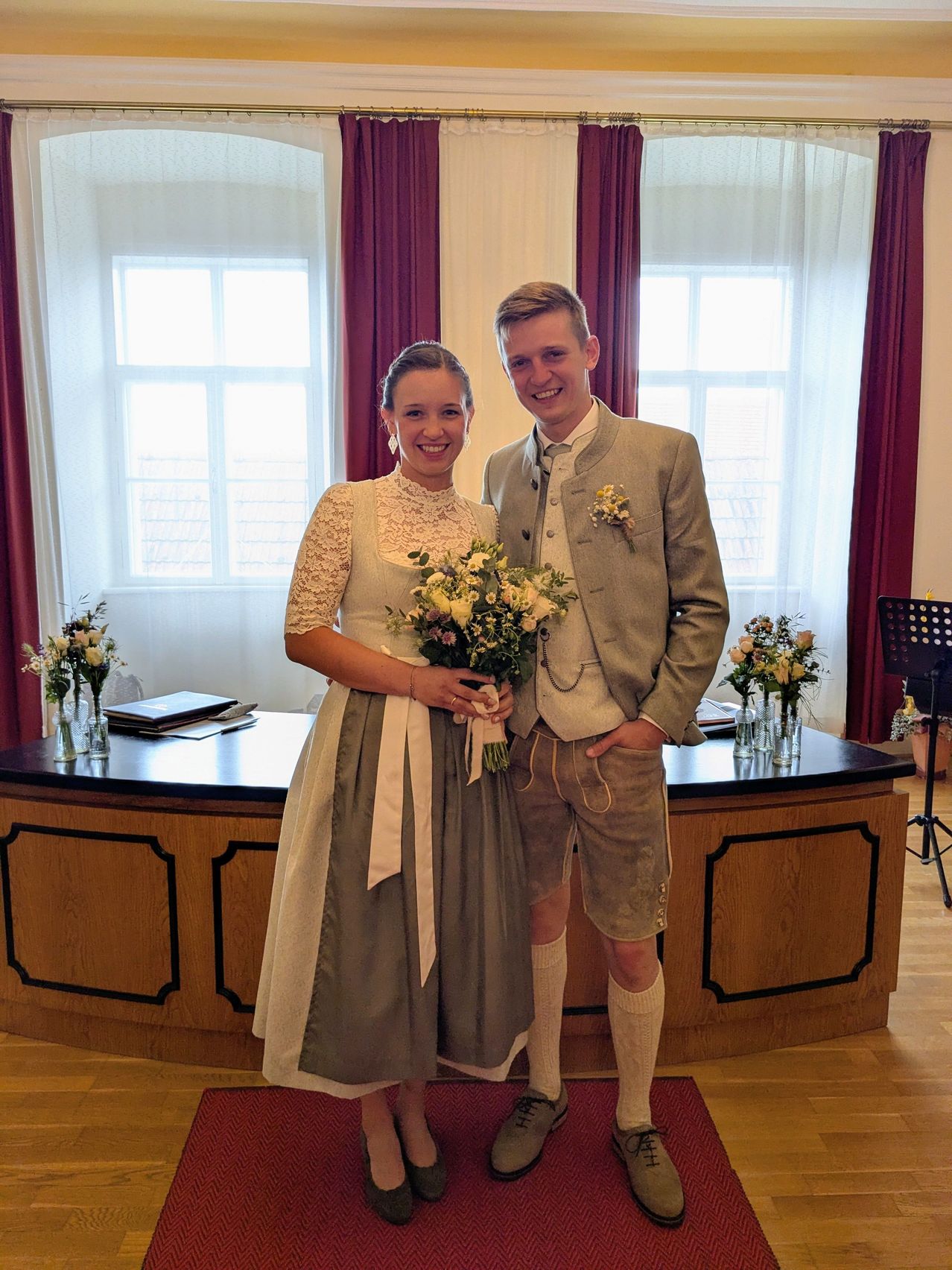 Ein lächelndes Paar steht vor einem Schreibtisch mit einem Blumenstrauß, gekleidet in traditioneller Kleidung, möglicherweise bei einer Hochzeitszeremonie.