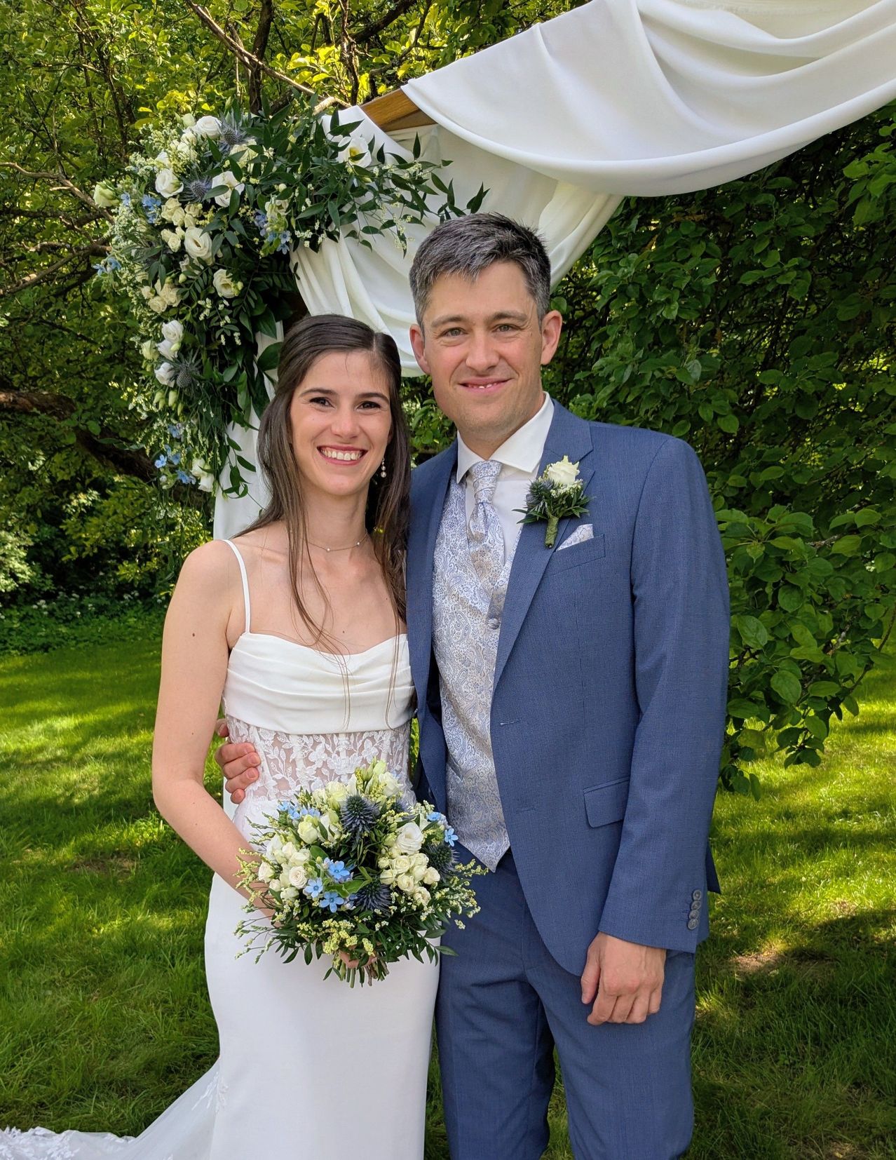 Ein lächelndes Paar steht unter einem Hochzeitsbogen, der Mann in einem blauen Anzug und die Frau in einem weißen Kleid mit einem Blumenstrauß.