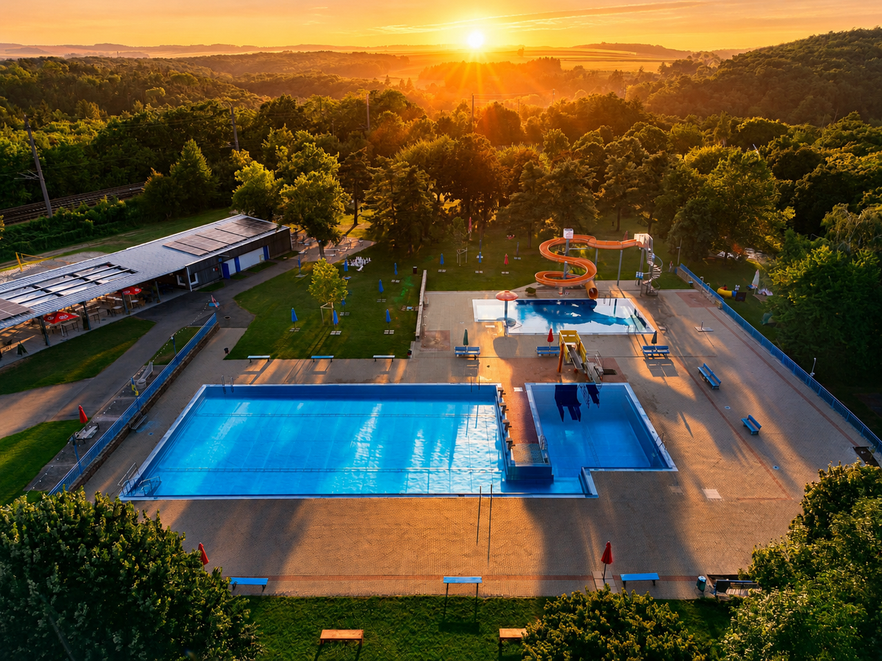 Eine Vogelperspektive eines Schwimmbadbereichs bei Sonnenuntergang mit einem großen Pool, einer Rutsche und einem kleineren Pool, umgeben von viel Grün und einem Gebäude mit Solarpanelen.