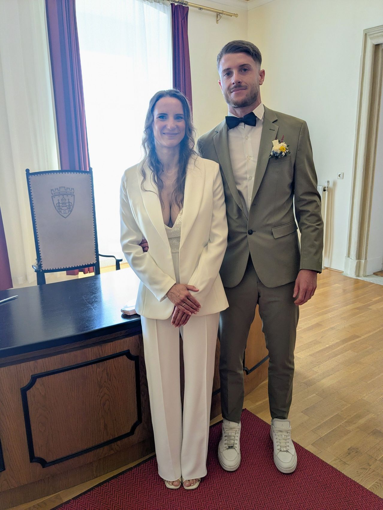 Ein Mann und eine Frau in formeller Kleidung stehen lächelnd nebeneinander, mit einem Holzschreibtisch und einem Stuhl im Hintergrund.