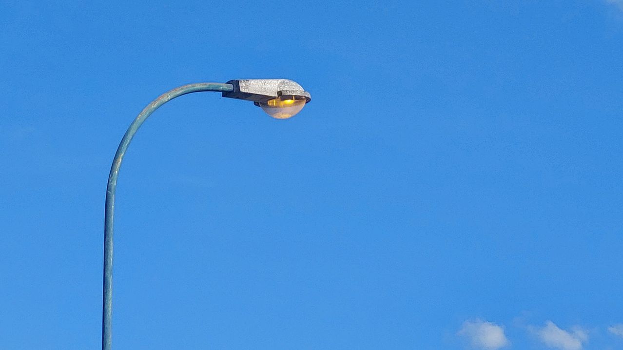 Eine Straßenlaterne ist an einem Pfosten gegen einen klaren blauen Himmel installiert. Das Licht ist an und die Glühbirne leuchtet gelb.