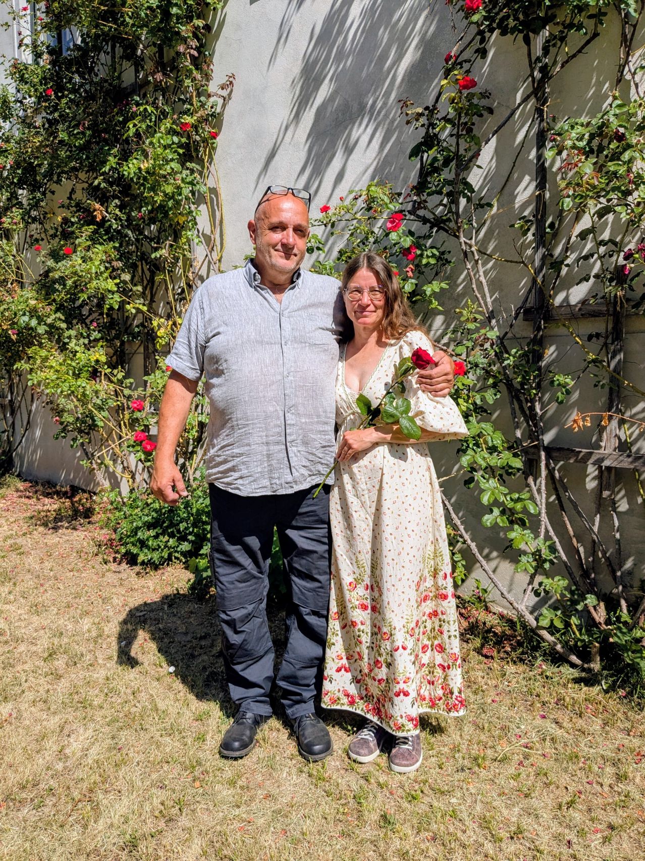 Ein Mann und eine Frau stehen zusammen in einem Garten, mit roten Blumen im Hintergrund. Der Mann trägt eine Brille und die Frau hält eine Rose.