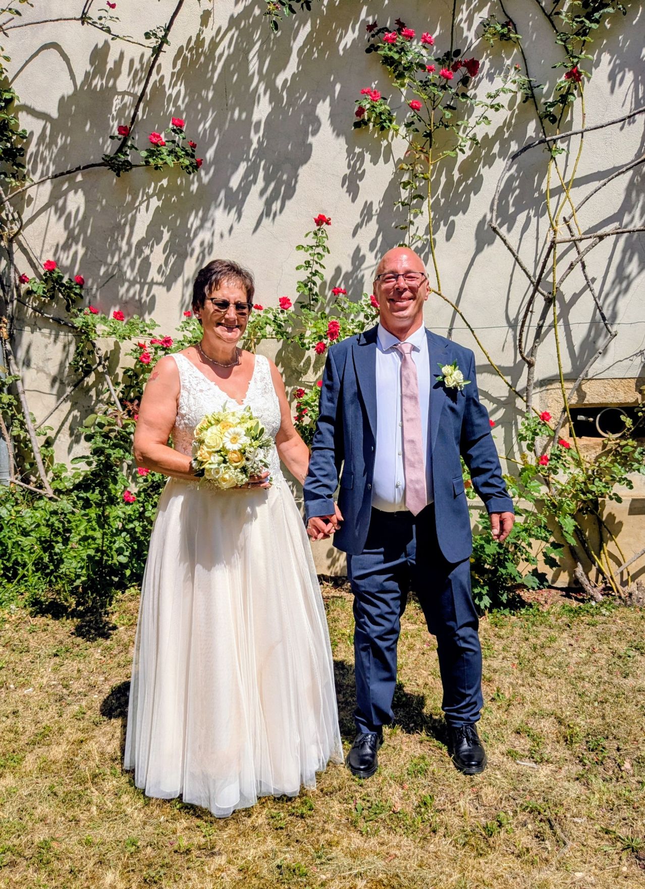 Eine lächelnde Braut in einem weißen Kleid hält einen Blumenstrauß und steht neben einem Bräutigam in einem blauen Anzug und einer rosa Krawatte in einem Garten mit roten Blumen und grünen Pflanzen.