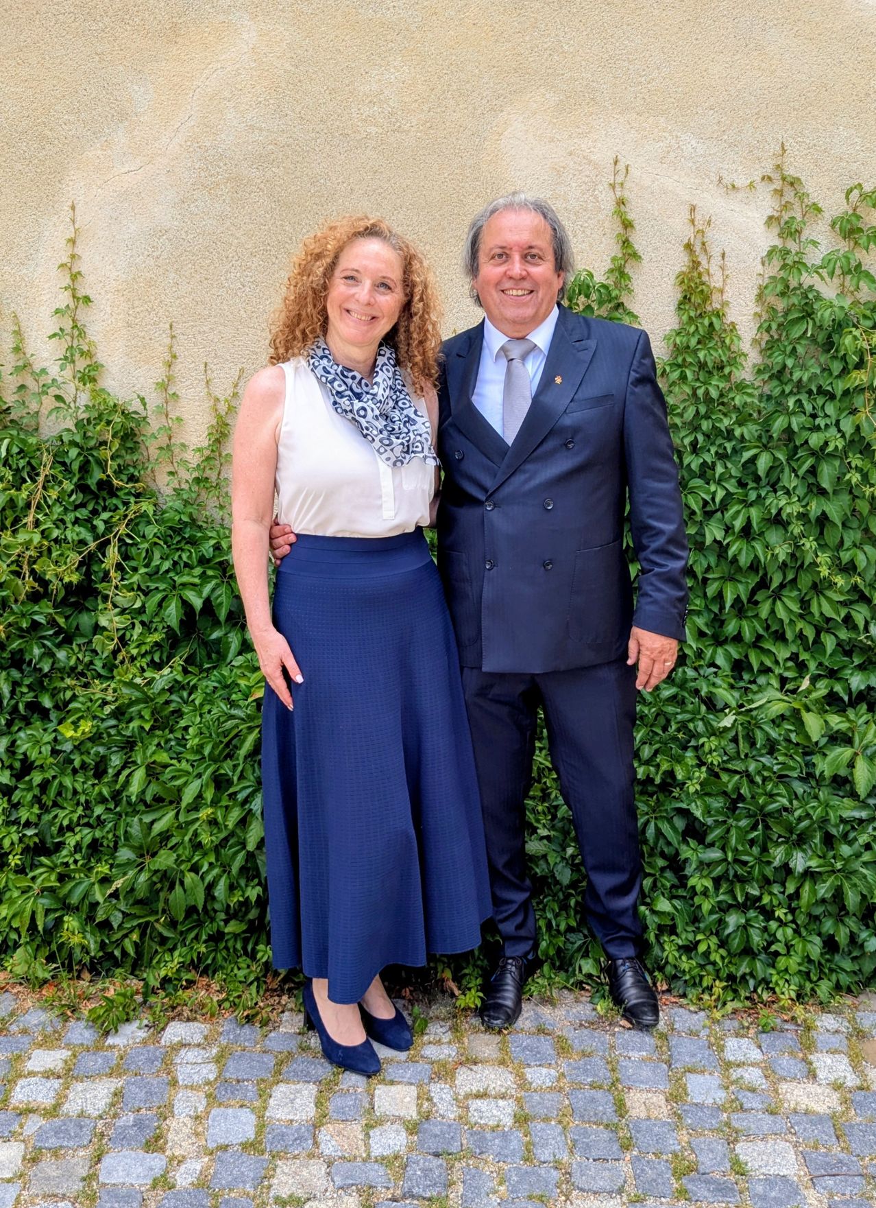Ein Mann und eine Frau stehen zusammen vor einer Wand mit grünem Laub. Die Frau trägt einen blauen Rock und ein weißes Oberteil. Der Mann trägt einen dunklen Anzug und eine Krawatte.