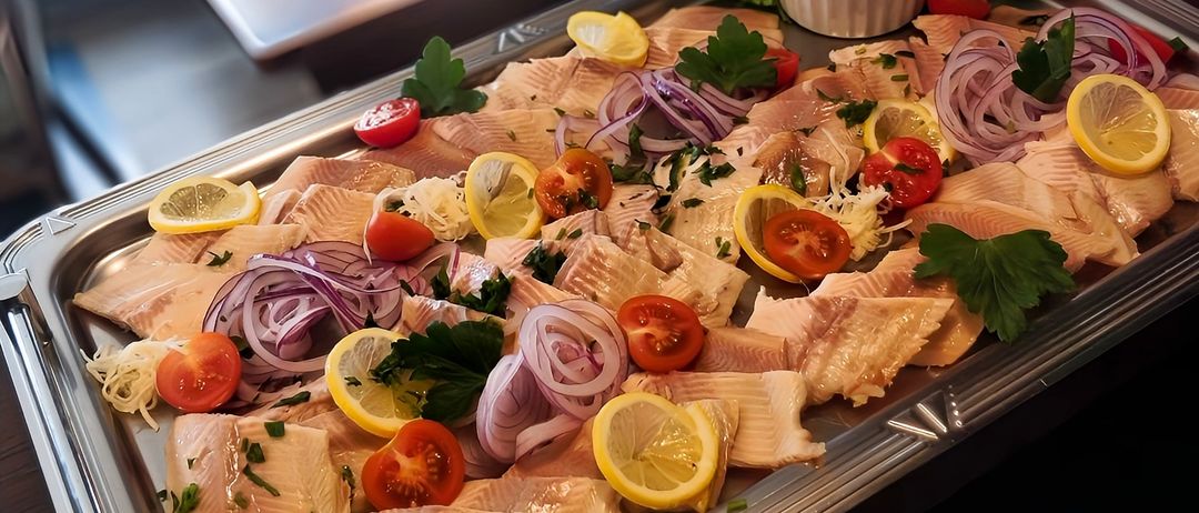 Ein Tablett mit geschnittenen Fischstücken, garniert mit Zitronen, Tomaten, Zwiebeln und Kräutern. Im Hintergrund ein Restaurant mit Tischen und Stühlen.