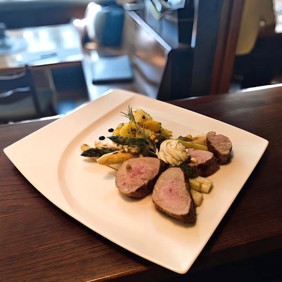Ein weißer Teller mit einem Gericht aus gebratenem Fleisch, Spargel und gelbem Gemüse auf einem Holztisch.