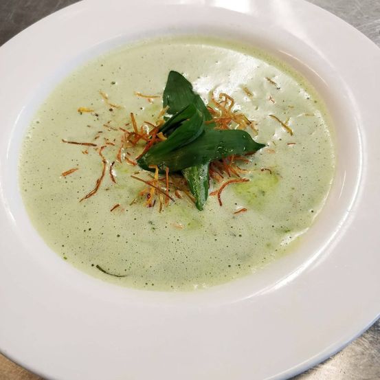 Ein weißer Teller mit einer cremigen grünen Suppe, garniert mit grünen Blättern und orangefarbener, geriebener Garnierung.