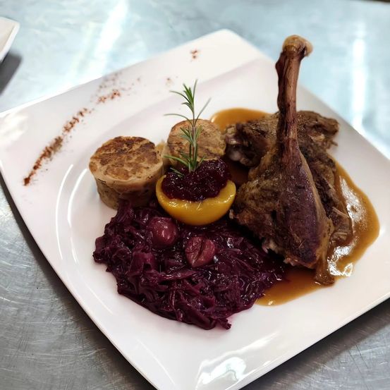 Ein weißer Teller mit einem Entenschenkel, rotem Kraut und einer gelben Frucht. Das Gericht ist mit einem Rosmarinzweig garniert.