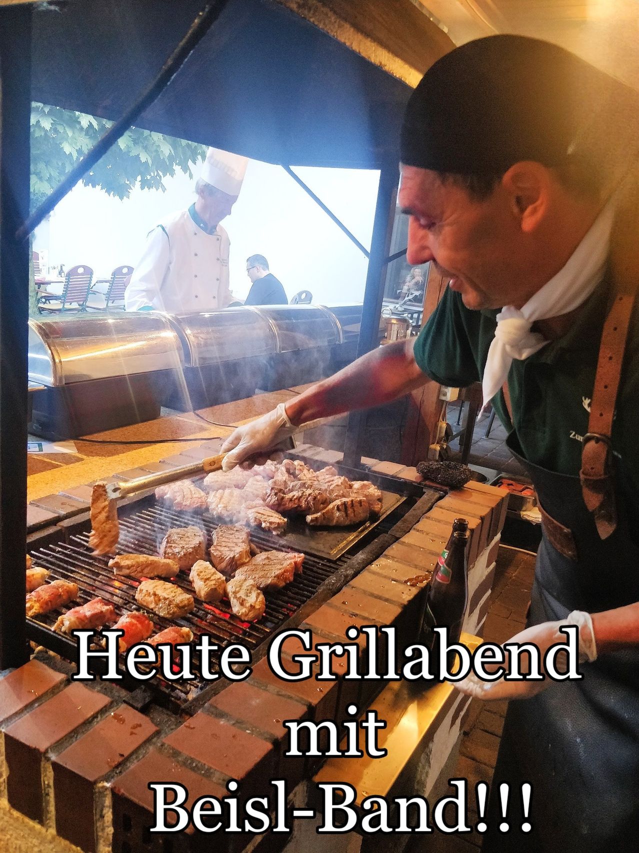 Ein Koch in einer Küche grillt Fleisch auf einem Grill mit einer großen Ziegelmauer. Ein anderer Koch steht hinter ihm. Neben dem Grill steht eine Flasche.