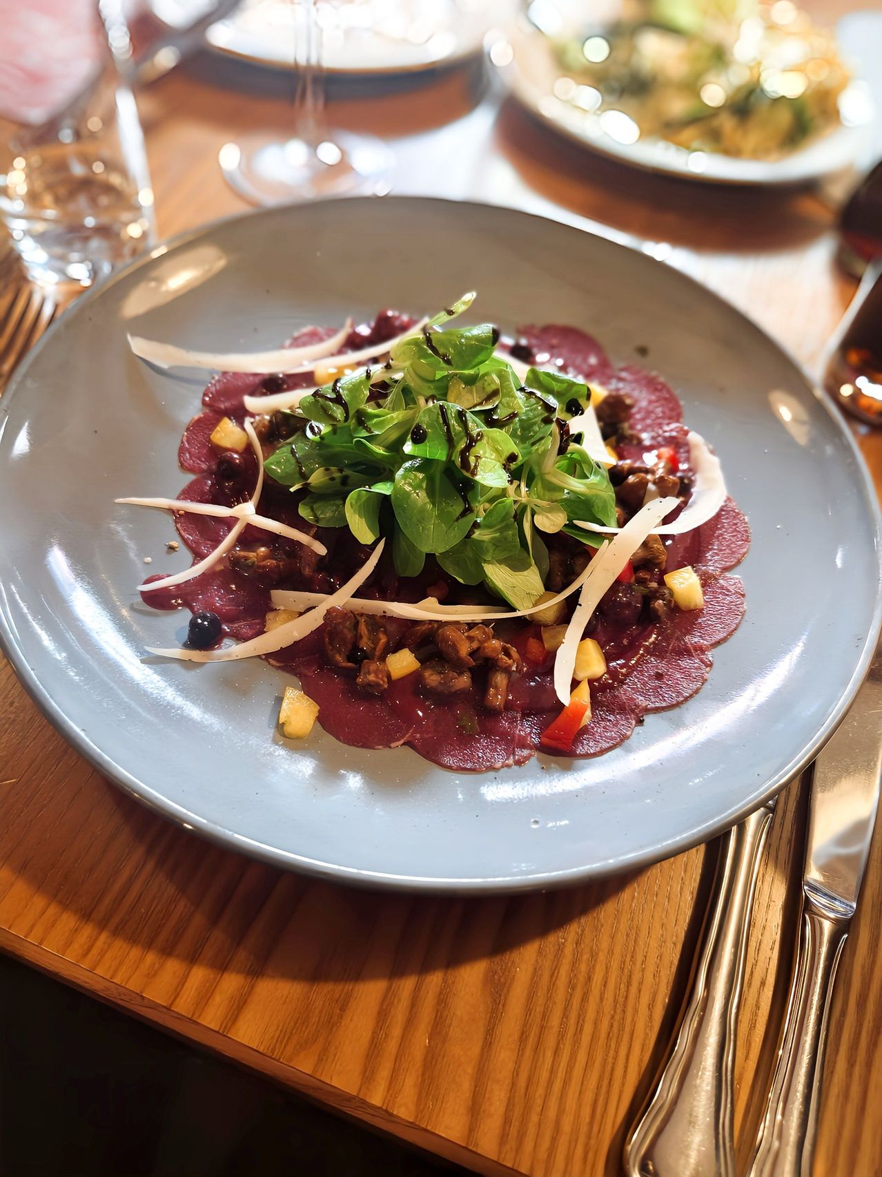 Ein Teller mit Fleisch, überzogen mit Salat, Zwiebeln und einer Sauce. Gabel, Messer und Glas auf dem Holztisch.