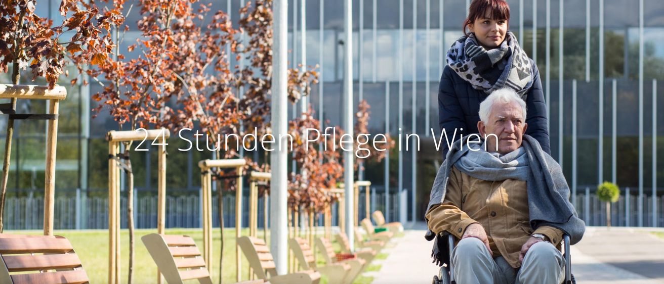 Ein Außenbereich mit mehreren Bänken, Bäumen und einem Gebäude mit Glaswänden. Ein Mensch im Rollstuhl und eine Frau mit einem Schal stehen in der Nähe.