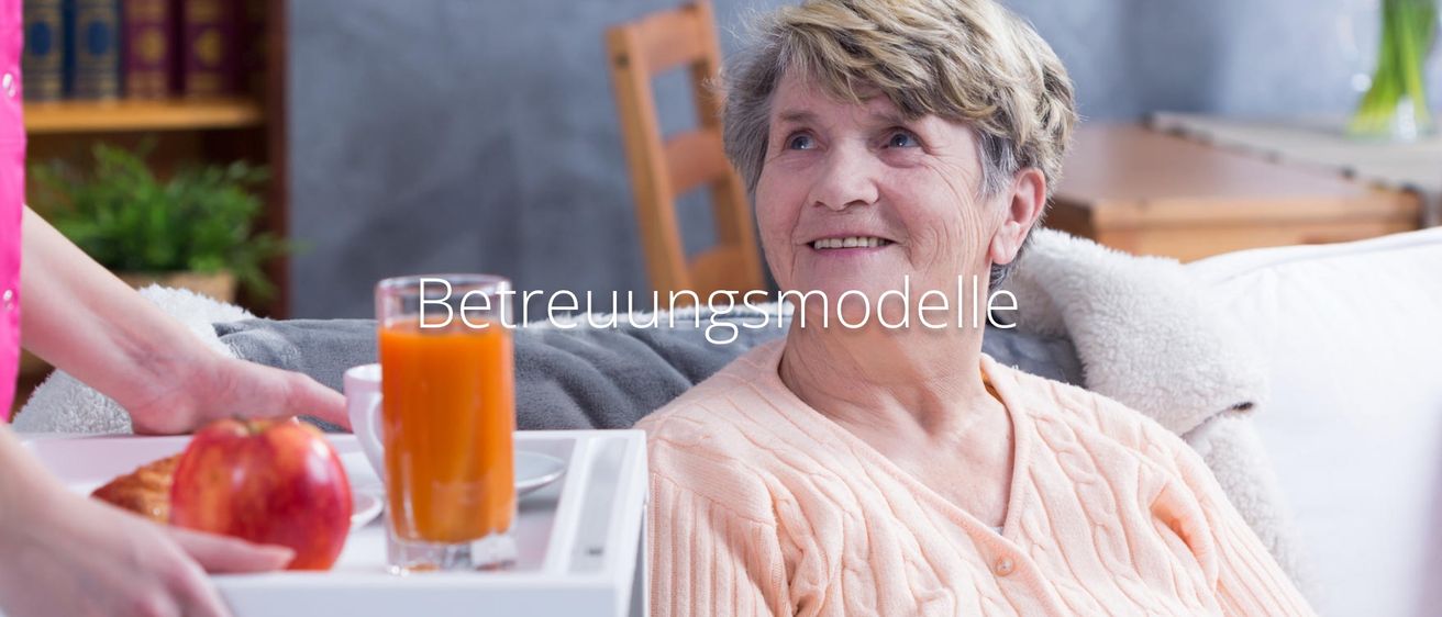 Eine ältere Frau mit kurzen blonden Haaren, die einen rosa Pullover trägt, sitzt auf einem Sofa, lächelt, mit einem Tablett in der Nähe. Hinter ihr ein Holzstuhl und ein Glas Orangensaft.