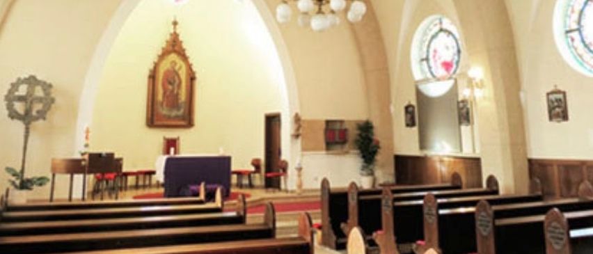 Ein leerer Kircheninnenraum mit gewölbten Decken, einer Kronleuchter und einem geschmückten Altar. Bänke sind auf beiden Seiten angeordnet. Eine Statue und ein Topfpflanzen im Hintergrund.