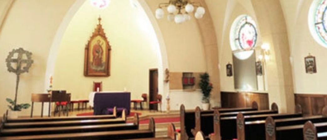 Eine leere Kirche mit Holzbänken und einem Altar mit einer Statue. Die Wände sind beige gestrichen und eine Laterne hängt von der Decke.