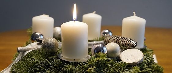Ein Kranz mit vier weißen Kerzen und silbernen Ornamenten steht auf einem hölzernern Tisch. Der Kranz besteht aus Tannennadeln und grünen Blättern. Die Kerzen sind angezündet und verbreiten ein warmes Licht.