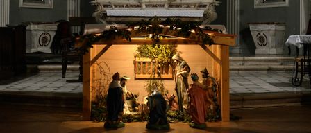 Eine Krippe ist in einer Kirche aufgestellt mit Statuen von Jesus, Maria und Josef sowie Tieren wie Esel und Lamm. Der Altar hat ein Kreuz und Blumen.