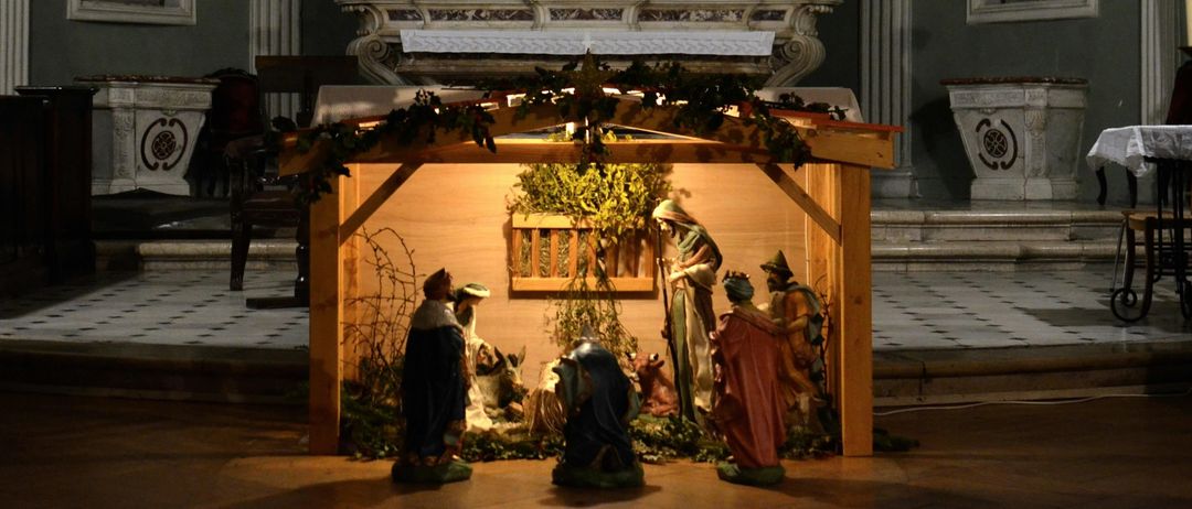 Eine Krippe ist vor einem Altar in einer Kirche aufgestellt. Sie enthält Statuen von Maria, Josef und Baby Jesus sowie Tiere. Die Szene ist von Grünpflanzen umgeben.
