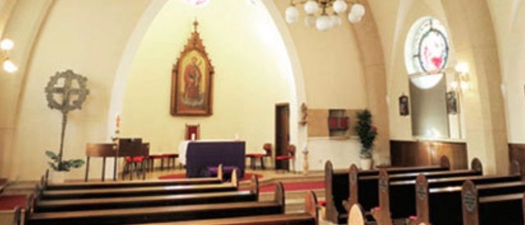 Ein Innenraum einer Kirche mit einem zentralen Altar, einer Laterne und Bänken auf beiden Seiten. Ein gerahmtes religiöses Bildnis ist an der Wand über dem Altar angebracht.