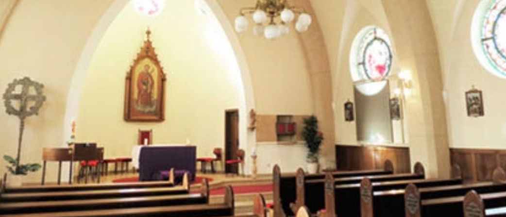 Das Innere einer Kirche mit gewölbten Decken und einer Kronleuchter. Bänke sind vor einem Altar mit einem religiösen Gemälde ausgerichtet.