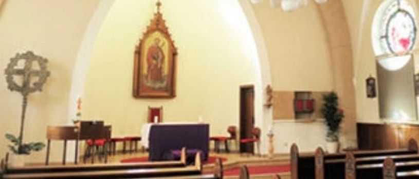 Ein Innenraum einer Kirche mit einer hängenden Kronleuchter. Der Altar ist mit einem Kreuz und einer religiösen Malerei dekoriert. Die Kirchenbänke sind in Reihen auf beiden Seiten angeordnet.