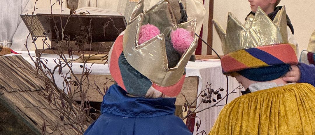 Ein Priester predigt bei einem Gottesdienst in einer Kirche, wobei Kinder in farbenfrohen Kronen und Umhängen aufmerksam zuhören.