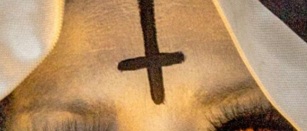 Eine Person ist als Nonne verkleidet, mit einem goldenen Kreuz auf der Stirn und roten Augen. Sie hält goldene Ketten und Ringe.