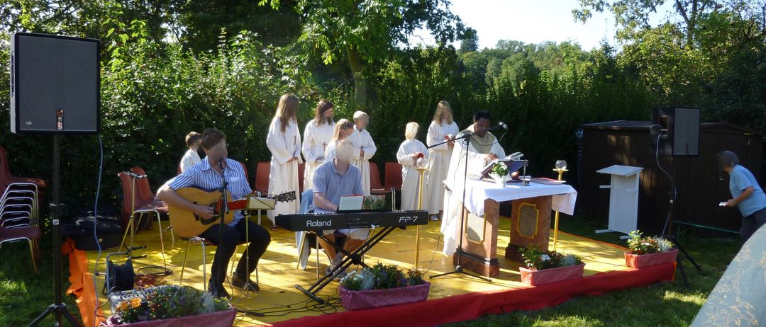 Eine Gruppe von Menschen in weißen Roben steht auf der Bühne in einem Park und führt eine Zeremonie durch. Eine Person spielt Gitarre, eine andere spielt Klavier. Ein Tisch mit einer Pflanze steht auf der Bühne. Bäume und Büsche umgeben den Bereich.