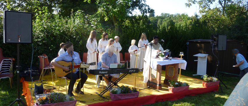 Eine Gruppe von Menschen in weißen Roben steht auf der Bühne in einem Park und führt eine Zeremonie durch. Eine Person spielt Gitarre, eine andere spielt Klavier. Ein Tisch mit einer Pflanze steht auf der Bühne. Bäume und Büsche umgeben den Bereich.
