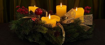 Ein Kranz aus immergrünen Pflanzen hält fünf gelbe Kerzen mit einem roten Apfel in der Mitte, umgeben von Tannenzapfen und roten Beeren, geschmückt mit goldenen Bändern auf einer schwarzen Oberfläche.