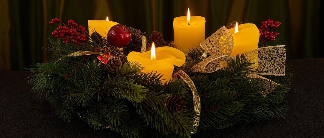 Ein Kranz aus immergrünen Pflanzen hält fünf gelbe Kerzen mit einem roten Apfel in der Mitte, umgeben von Tannenzapfen und roten Beeren, geschmückt mit goldenen Bändern auf einer schwarzen Oberfläche.