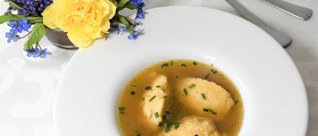 Eine Schüssel Suppe mit Knödeln und Kräutern, auf einem Tisch mit Blumen und Löffeln.