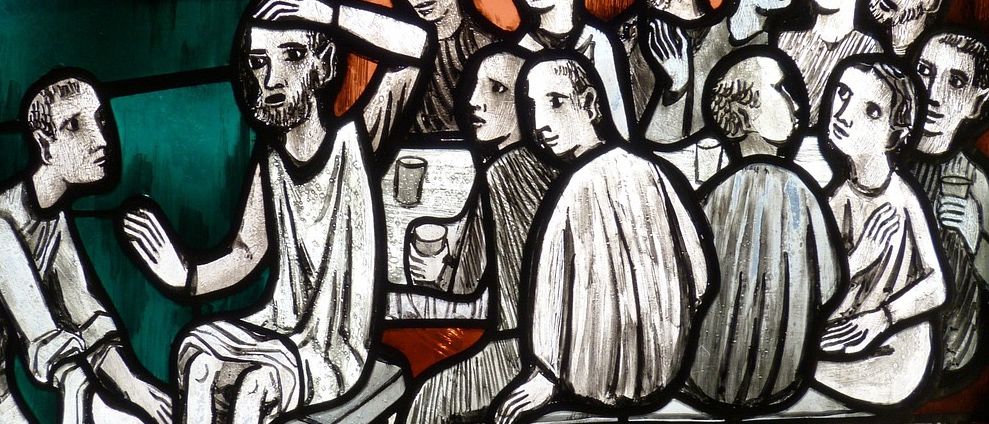 Ein Buntglasbild zeigt eine Szene aus biblischen Zeiten, bei der Jesus einem Jünger die Füße wäscht, während viele andere um ihn herum sitzen.