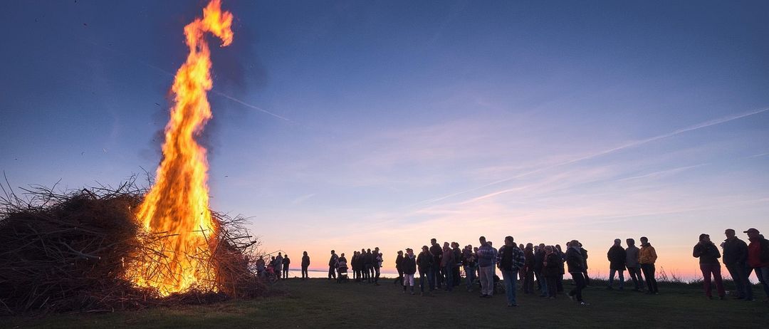 Ein großes Lagerfeuer brennt hell auf einem Hügel, während sich Menschen um es herum versammeln und unter einem klaren, dämmrigen Himmel zusehen.