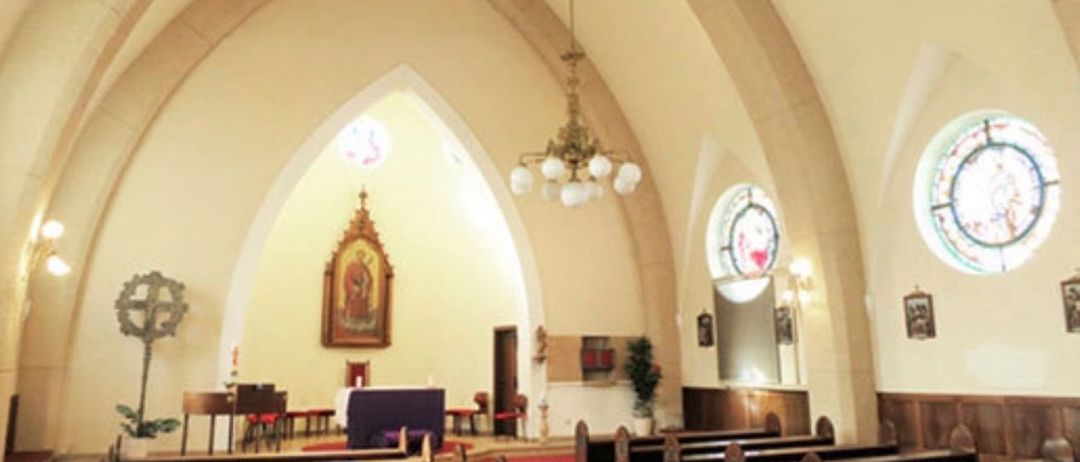 Der leere Kircheninnenraum zeigt einen Altar mit einer religiösen Ikone, einer Kronleuchter und Buntglasfenster. Bänke sind auf beiden Seiten des Ganges angeordnet.