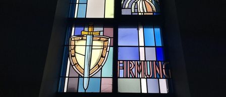 A stained glass window displays a sword and shield design with colorful tiles and the word 'Firmung' below it.