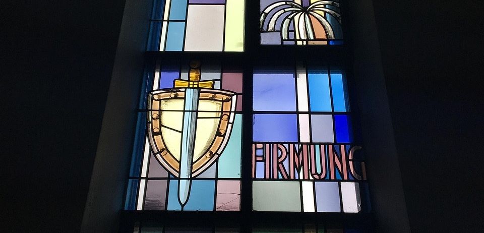Ein Buntglasfenster zeigt ein Schwert- und Schilddesign mit farbigen Kacheln und dem Wort 'Firmung' darunter.