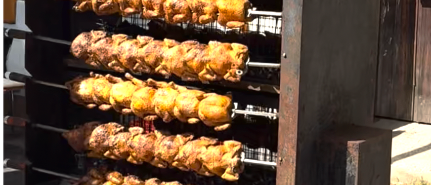 Ein Nahaufnahme eines Grills mit mehreren Hähnchenspießen. Der Grill ist aus Edelstahl mit einem geneigten Dach. Das Essen scheint gut gegart zu sein.