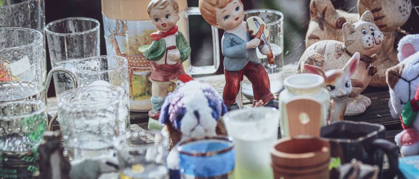 Eine Sammlung von Glasbechern, Tassen und Figuren ist auf einem Holztisch ausgestellt. Die Figuren umfassen einen Jungen, der eine Geige spielt, ein Mädchen, das ein Buch hält, und einen Hund.