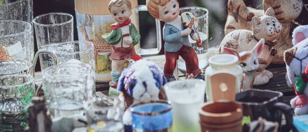 Eine Sammlung von Glasbechern, Tassen und Figuren ist auf einem Holztisch ausgestellt. Die Figuren umfassen einen Jungen, der eine Geige spielt, ein Mädchen, das ein Buch hält, und einen Hund.