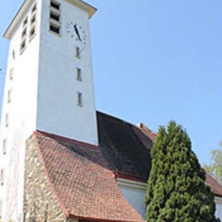 Eine weiße Kirche mit einem braunen Dach und einem hohen Turm, der eine Uhr zeigt. Bäume und Pflanzen umgeben das Gebäude.