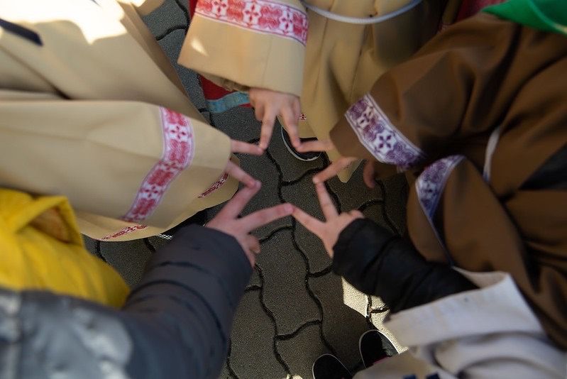 Mehrere Kinder in traditioneller Kleidung machen ein Friedenszeichen mit ihren Fingern. Sie stehen im Kreis auf einem Betonboden.