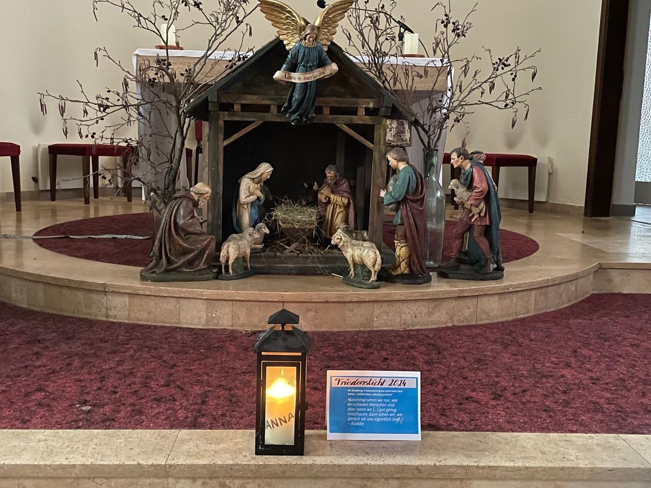 Eine Krippe ist in einer Kirche aufgestellt und zeigt Statuen von Maria, Josef und Jesus sowie Hirten und Schafe. Ein beleuchteter Laternen mit dem Wort Anna und einem blauen Schild mit Text ist in der Nähe.