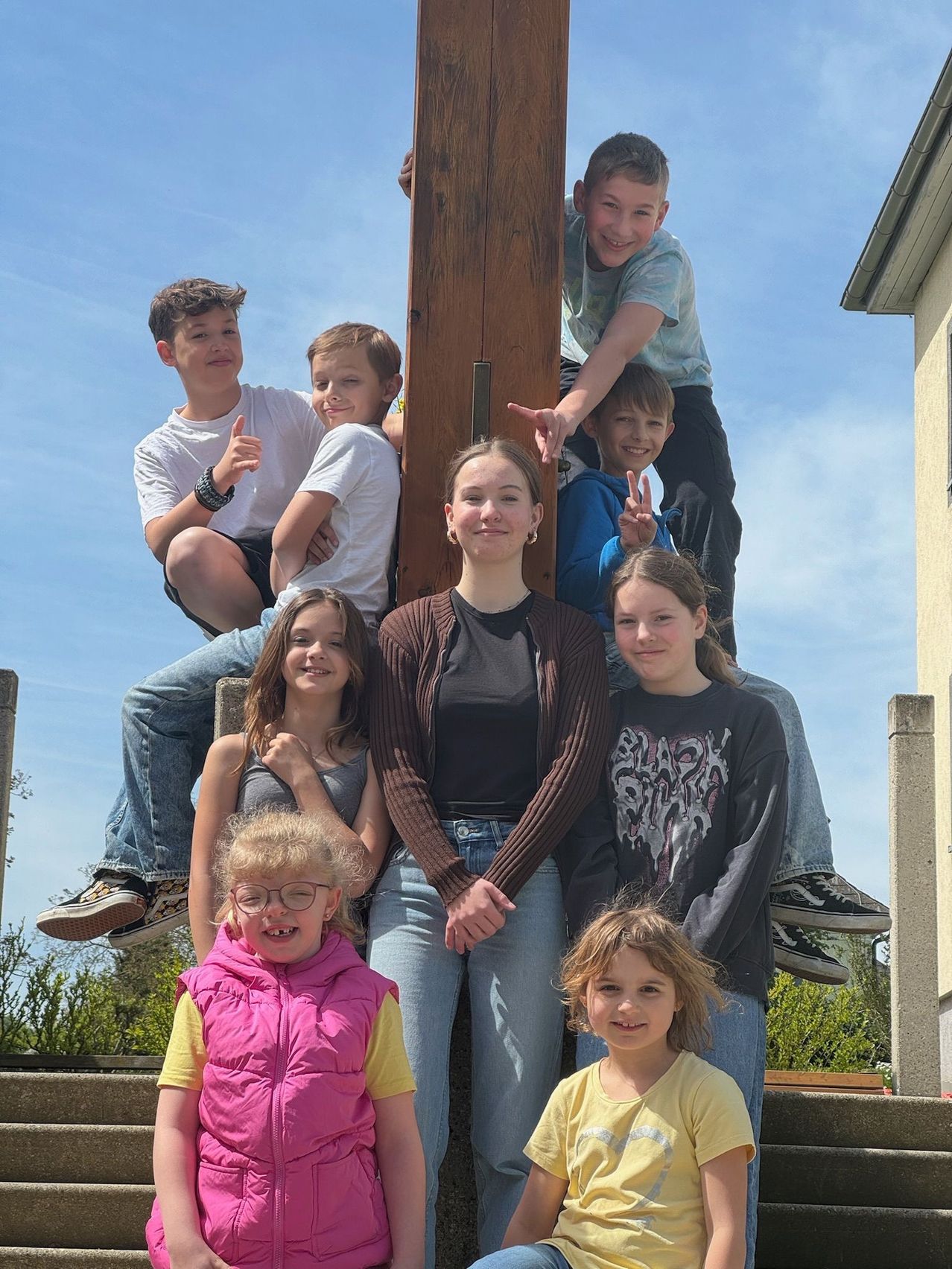 Eine Gruppe von Kindern und Jugendlichen posiert für ein Foto im Freien. Einige stehen, andere sitzen auf der Kante eines Holzpfostens. Sie lächeln und schauen zur Kamera.